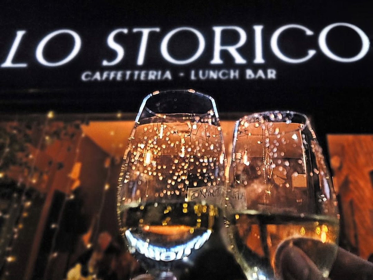 Due calici di vino bianco frizzante in un brindisi sotto l'insegna Lo Storico Caffetteria - Lunch Bar, luci calde sfocate