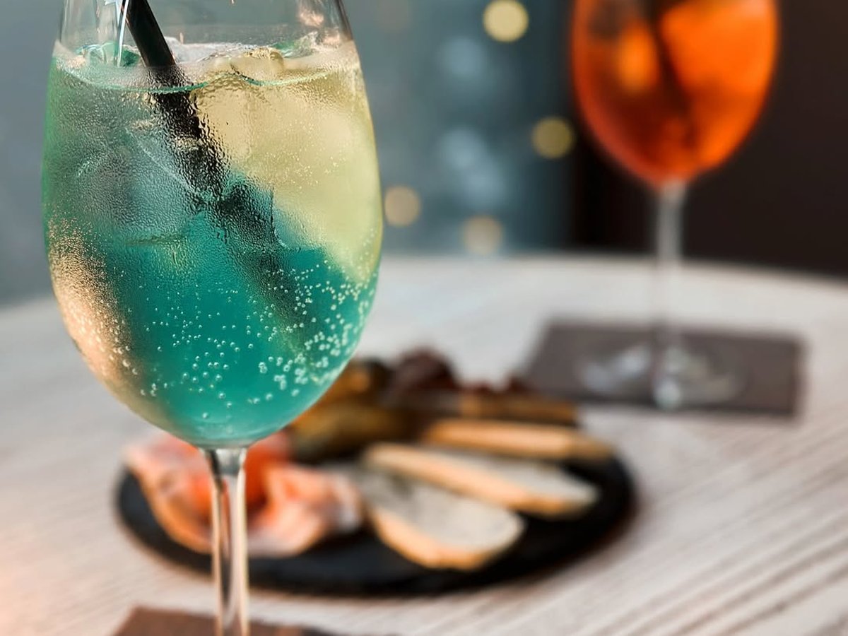 Calice di cocktail azzurro con ghiaccio e cannuccia nera, spritz arancione e tagliere di ardesia con salumi e crostini