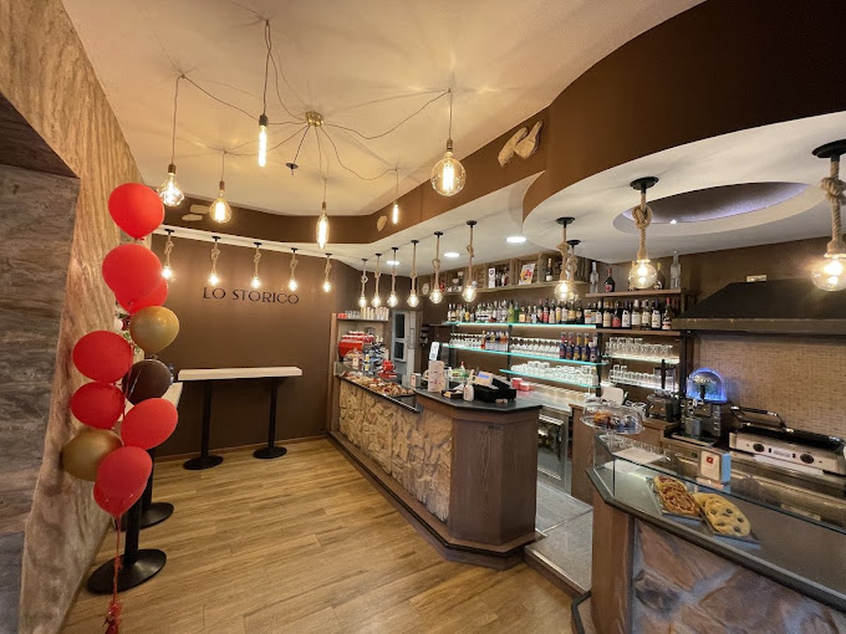 Bancone in pietra del bar Lo Storico con lampadine pendenti, bottiglie sugli scaffali e palloncini rossi e oro